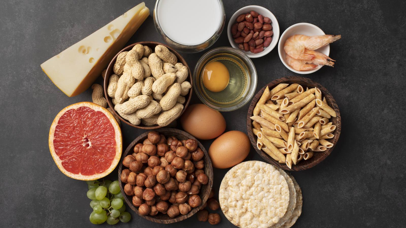 Mesa escura com alimentos que contêm alergénios comuns: queijo, leite, amendoins, ovo, camarão, massa, avelãs e sementes de sésamo.